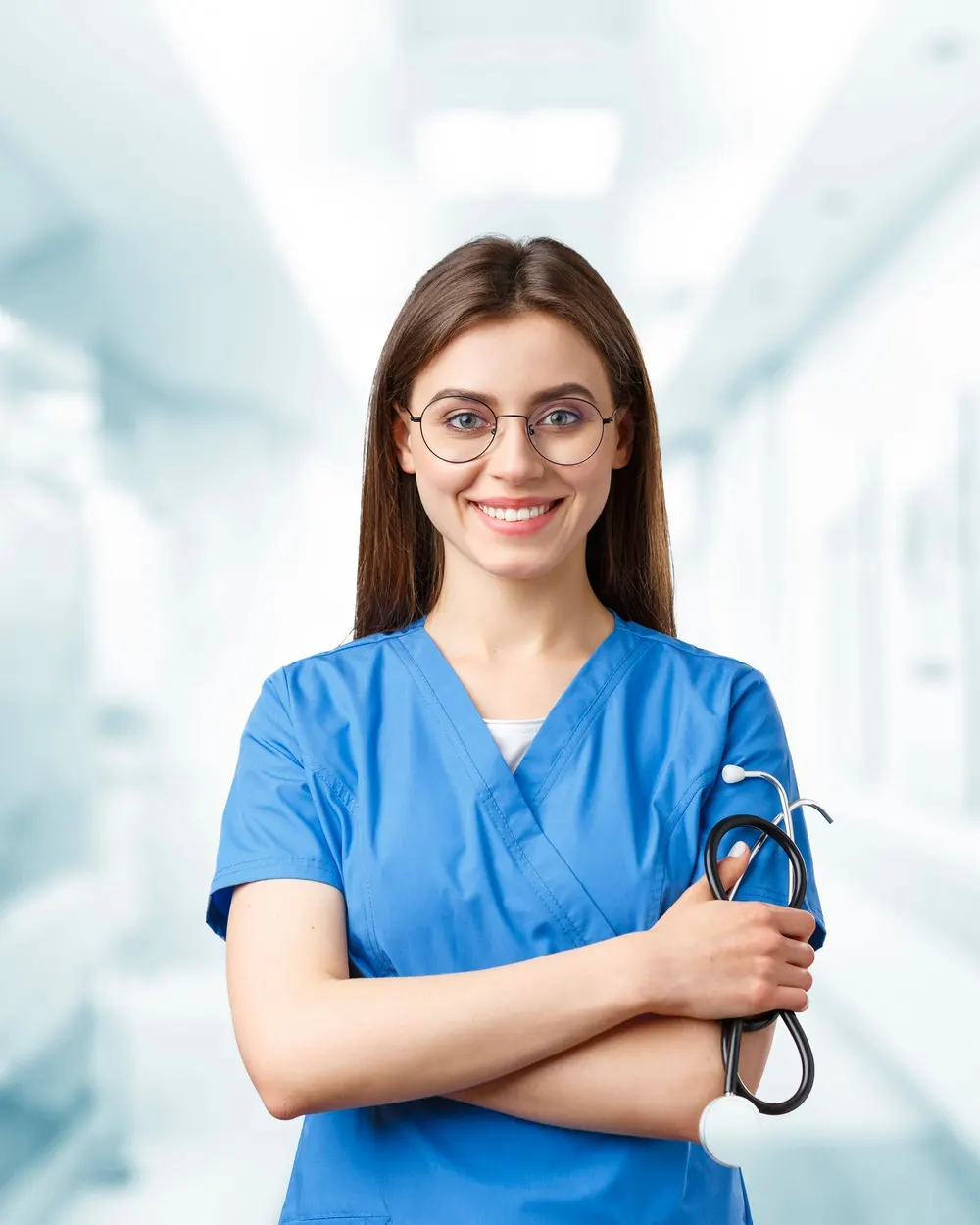 Smiling nurse in blue scrubs holding a stethoscope in a hospital corridor